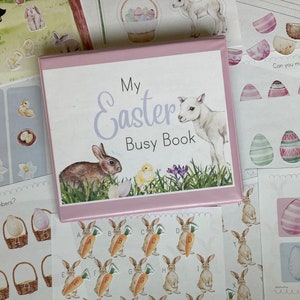 以下が含まれることがあります： 表紙に「My Easter Busy Book」というタイトルが書かれたピンク色の本。表紙には、茶色のウサギ、白い子羊、黄色いひよこが描かれています。この本は、卵のマッチング、ひよこの数え上げ、数字のマッチングなど、さまざまなイースターをテーマにしたアクティビティで囲まれています。