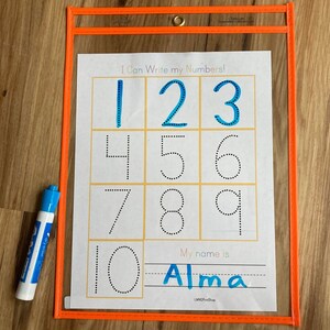 May include: A clear plastic sheet with a white sheet inside. The white sheet has a title that says "I Can Write My Numbers!" and has dotted outlines of the numbers 1 through 10. There is a line below the numbers that says "My name is" and a line for writing a name. The name "Alma" is written in blue on the line. A blue dry-erase marker is next to the sheet.