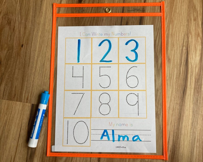 May include: A clear plastic sheet with a white sheet inside. The white sheet has a title that says "I Can Write My Numbers!" and has dotted outlines of the numbers 1 through 10. There is a line below the numbers that says "My name is" and a line for writing a name. The name "Alma" is written in blue on the line. A blue dry-erase marker is next to the sheet.