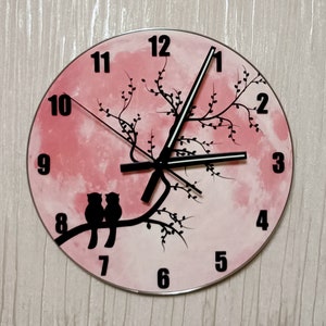 Op de afbeelding: Ronde wandklok met een roze achtergrond en een zwart takontwerp. Twee zwarte uil silhouetten zitten op de tak. Zwarte cijfers markeren de uren. De klok heeft zwarte wijzers en een zilveren metalen frame.
