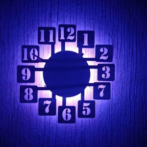 Può includere: Un orologio in metallo nero con numeri bianchi su una parete blu. L'orologio è illuminato da dietro, creando un effetto di bagliore.