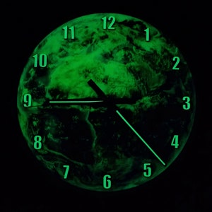 Puede incluir: Un reloj que brilla en la oscuridad con una imagen de la Tierra verde y negra. La esfera del reloj tiene los números del 1 al 12 y las manecillas son negras.