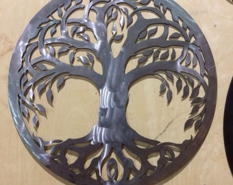 May include: A circular metal wall art piece depicting a tree of life design. The tree has a thick trunk and branches with leaves, and intricate roots. The metal is brushed and has a silver finish.