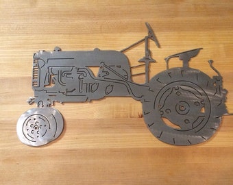 May include: A metal silhouette of a vintage tractor with detailed engine and wheel designs. The tractor is cut out of metal and mounted on a wooden surface.