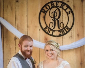 May include: A bride and groom cutting a white wedding cake with a wooden topper that says "Roberts" and features a couple and a dog. The cake is on a white cake stand and there are cupcakes on tiered stands on either side of the cake. The couple is standing in front of a wooden wall with a white fabric backdrop and a black metal sign that says "Roberts Est. 2018" with a cursive "R" in the center.