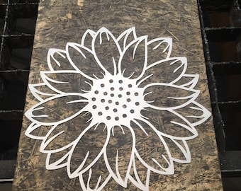 May include: A white metal sunflower cutout on a brown wooden surface. The sunflower has a detailed design with petals and a center.