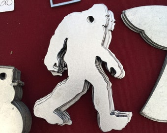 May include: A white metal cutout of a Bigfoot walking, layered on top of itself. The cutout is on a red background with other metal cutouts, including a snowman and the letter "M".