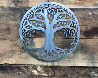 May include: A silver metal tree of life wall decor with intricate branches and roots. The tree is cut out of a circular piece of metal and has a rustic, weathered look.