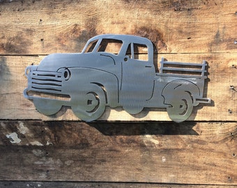 May include: A metal silhouette of a vintage pickup truck, mounted on a wooden wall. The truck is a classic design with a flatbed and a simple grille.