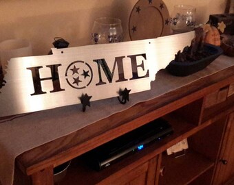 May include: A silver metal cutout of the state of Tennessee with the word "HOME" and three stars in a circle in the center. The cutout is mounted on a wooden shelf with a brown runner.