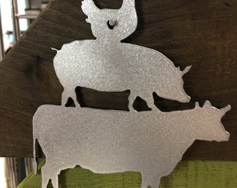 May include: A metal silhouette of a cow, pig, and chicken stacked on top of each other. The silhouettes are silver and are mounted on a wooden background.