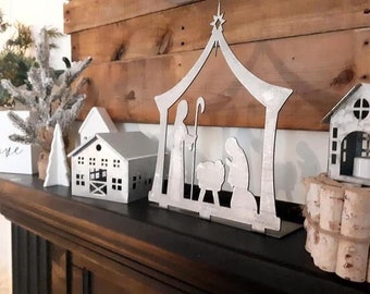 May include: A silver metal nativity scene with a cutout of Mary, Joseph, and the baby Jesus in a manger. The scene is on a wooden shelf with a white sign that says "love" and other decorative items.