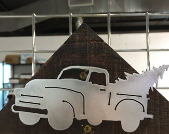 May include: A metal cutout of a vintage pickup truck with a Christmas tree in the bed, mounted on a wooden triangle. The truck is silver and the wood is brown.