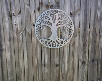 May include: A silver metal tree of life wall decor with a circular design. The tree has a thick trunk and branches with leaves. The decor is mounted on a wooden fence.
