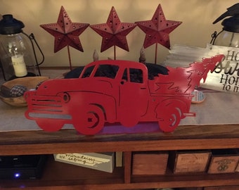 May include: Red metal cutout of a pickup truck carrying a Christmas tree. The truck is on a wooden shelf with other decorations, including three red metal stars.