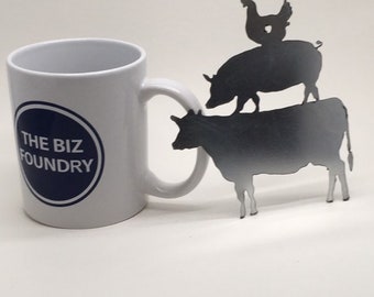 May include: White ceramic mug with a blue circle logo that says "THE BIZ FOUNDRY". A metal silhouette of a cow, pig, and chicken is behind the mug.