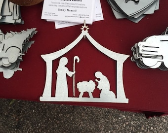 May include: White metal cutout of a nativity scene with a star on top. The scene includes Mary, Joseph, and the baby Jesus in a manger with a sheep. The cutout is on a red surface.
