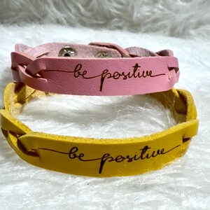 Könnte beinhalten: Zwei Lederarmbänder, eines rosa und eines gelb, jeweils mit dem kursiv geschriebenen Schriftzug "be positive". Das rosa Armband hat ein geflochtenes Design und einen Druckknopfverschluss. Das gelbe Armband hat ein gewebtes Design.
