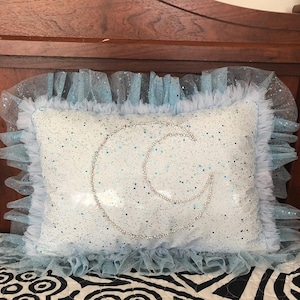 May include: A light blue pillow with a crescent moon design made of white glitter. The pillow has a light blue tulle ruffle around the edges.