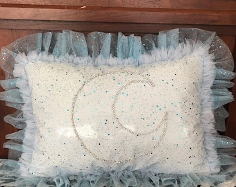 May include: A light blue pillow with a crescent moon design made of white glitter. The pillow has a light blue tulle ruffle around the edges.