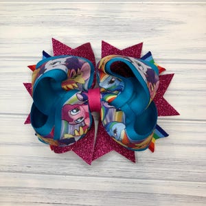 May include: A large, handmade hair bow featuring a teal ribbon with a cartoon character print and a hot pink center. The bow is accented with glittery, magenta starburst shapes.