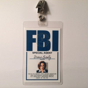 May include: A white plastic ID card with a blue FBI logo and the text "SPECIAL AGENT Dana Scully" with a photo of a woman in a white shirt. The text "THIS IS TO CERTIFY THAT WHOSE SIGNATURE AND PHOTOGRAPH APPEARS HEREON IS A REGULARLY APPOINTED SPECIAL AGENT OF THE FBI" is printed below the photo.