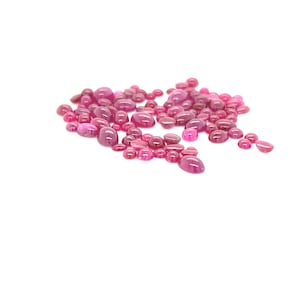 Puede incluir: Una colección de cabujones de rubí rosa de varios tamaños, esparcidos sobre un fondo blanco.