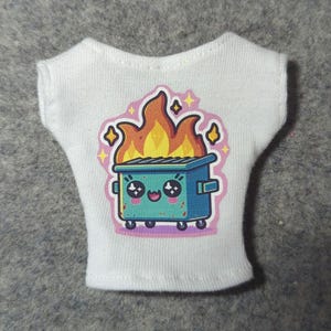 May include: White sleeveless shirt featuring a colorful graphic of a blue dumpster on fire with a cute face. The flames are orange and yellow, and the design includes pink and yellow accents. The shirt is made of a soft, white fabric.