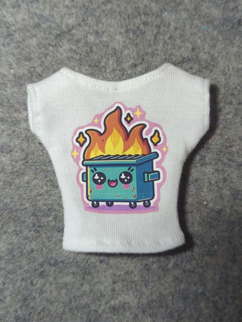 May include: White sleeveless shirt featuring a colorful graphic of a blue dumpster on fire with a cute face. The flames are orange and yellow, and the design includes pink and yellow accents. The shirt is made of a soft, white fabric.