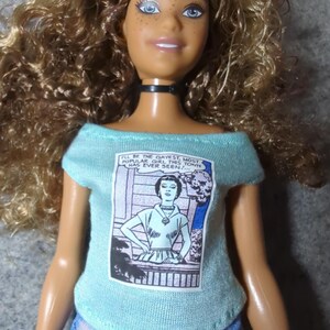 May include: A doll with curly brown hair and blue eyes wearing a light blue off-the-shoulder top and denim jeans. The top features a comic book-style graphic with the text "I'LL BE THE GAYEST, MOST POPULAR GIRL THIS TOWN HAS EVER SEEN!"