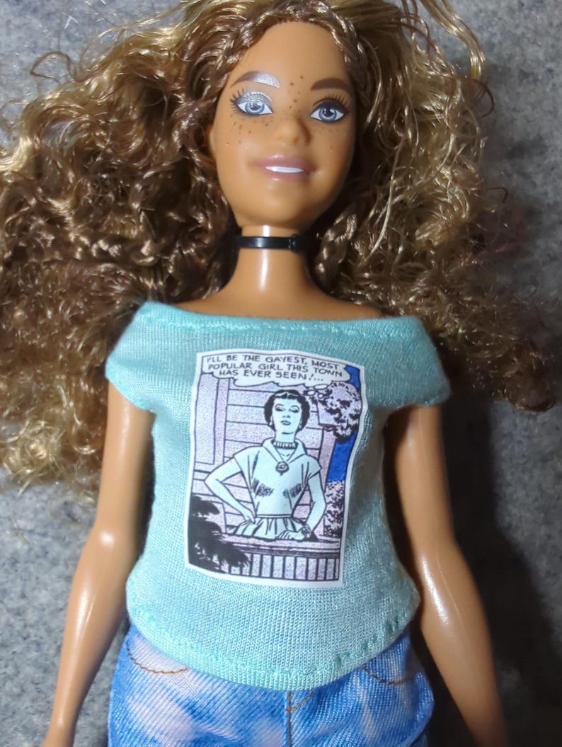 May include: A doll with curly brown hair and blue eyes wearing a light blue off-the-shoulder top and denim jeans. The top features a comic book-style graphic with the text "I'LL BE THE GAYEST, MOST POPULAR GIRL THIS TOWN HAS EVER SEEN!"
