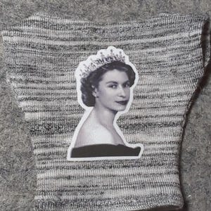 May include: A gray and white knitted fabric with a black and white sticker of a woman wearing a crown. The sticker is of a woman with dark hair and a dark top.