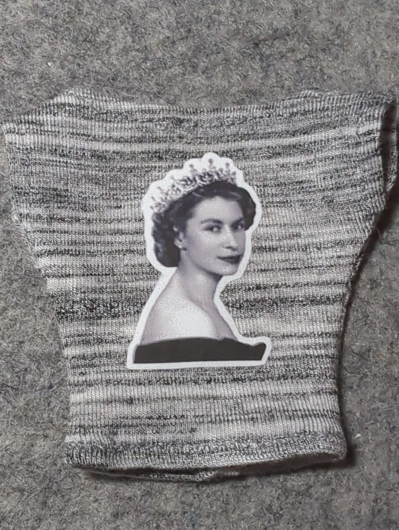 May include: A gray and white knitted fabric with a black and white sticker of a woman wearing a crown. The sticker is of a woman with dark hair and a dark top.