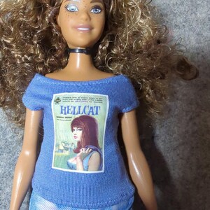 May include: A doll with long, curly brown hair, blue eyes, and freckles, wearing a blue t-shirt with a "HELLCAT" magazine cover graphic and tie-dye shorts. The doll has a black choker necklace.