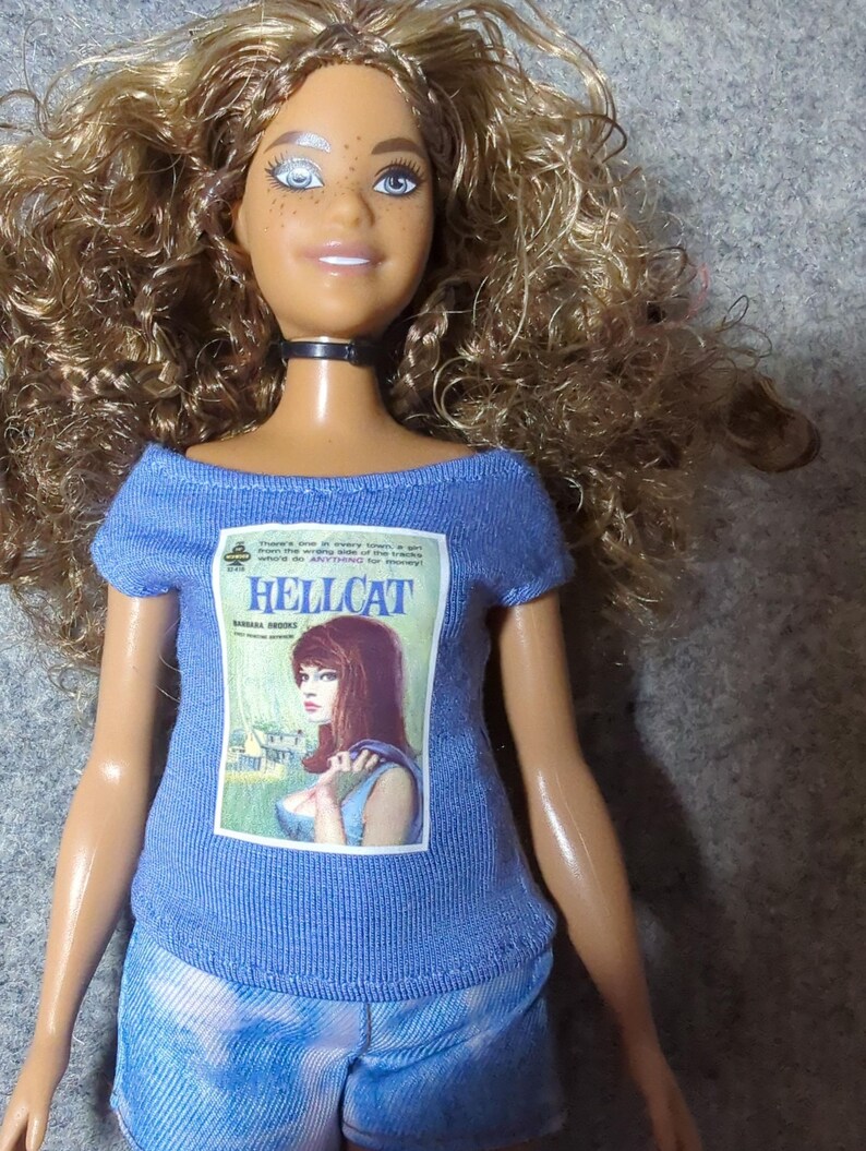 May include: A doll with long, curly brown hair, blue eyes, and freckles, wearing a blue t-shirt with a "HELLCAT" magazine cover graphic and tie-dye shorts. The doll has a black choker necklace.