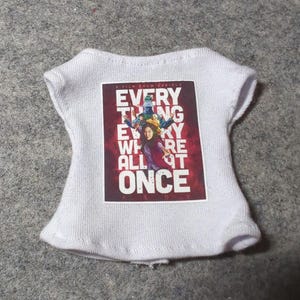 May include: White knit top with a rectangular graphic featuring the text "EVERYTHING EVERYWHERE ALL AT ONCE" in a bold, stylized font. The graphic has a red background with images of people.