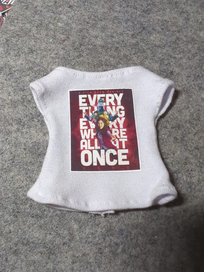 May include: White knit top with a rectangular graphic featuring the text "EVERYTHING EVERYWHERE ALL AT ONCE" in a bold, stylized font. The graphic has a red background with images of people.