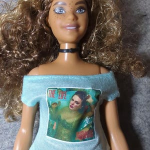 May include: A doll with light brown curly hair, blue eyes, and freckles, wearing a black choker and a light blue off-the-shoulder shirt. The shirt features a graphic with the text "THE LIFE" and an image of a woman.