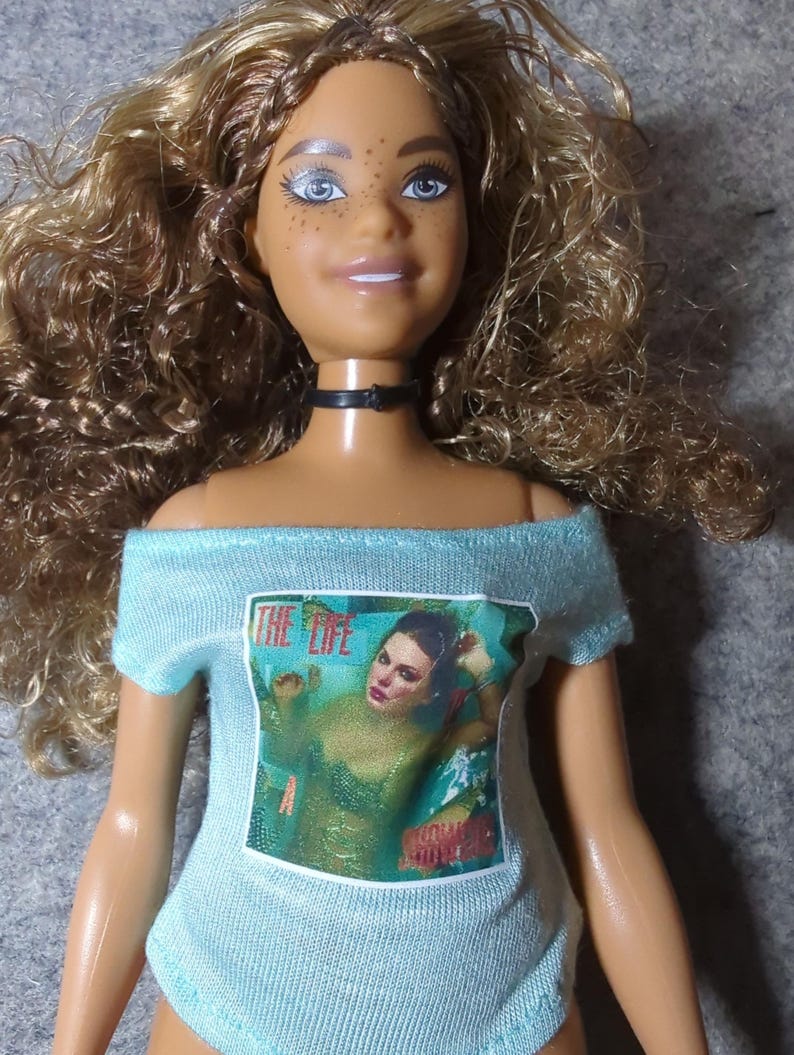 May include: A doll with light brown curly hair, blue eyes, and freckles, wearing a black choker and a light blue off-the-shoulder shirt. The shirt features a graphic with the text "THE LIFE" and an image of a woman.