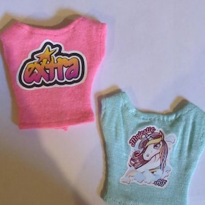 May include: Two doll-sized t-shirts. One is pink with the word "EXTRA" in a bubble-style font. The other is light blue with a unicorn graphic and the word "Majestic". Both shirts have a scoop neck.