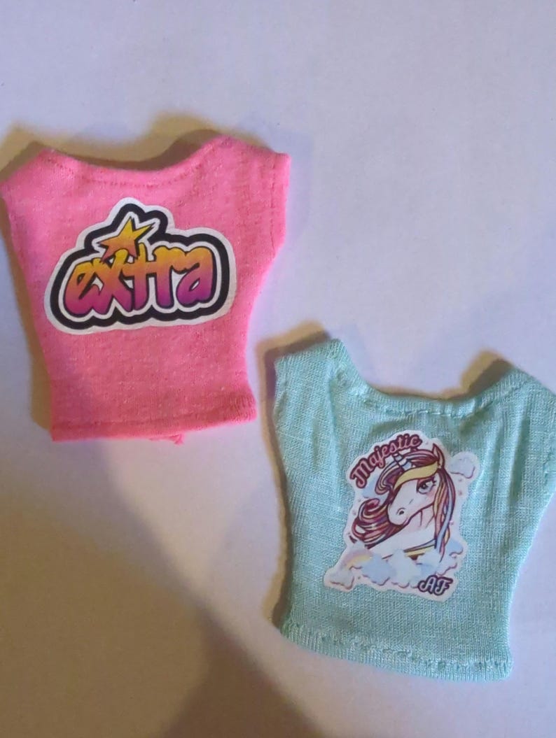 May include: Two doll-sized t-shirts. One is pink with the word "EXTRA" in a bubble-style font. The other is light blue with a unicorn graphic and the word "Majestic". Both shirts have a scoop neck.