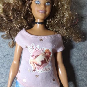 May include: A doll with curly blonde hair, blue eyes, and freckles, wearing a pink off-the-shoulder t-shirt with the words "Short n' Sweet" and a graphic of a woman. The doll is also wearing blue denim shorts and a black choker.