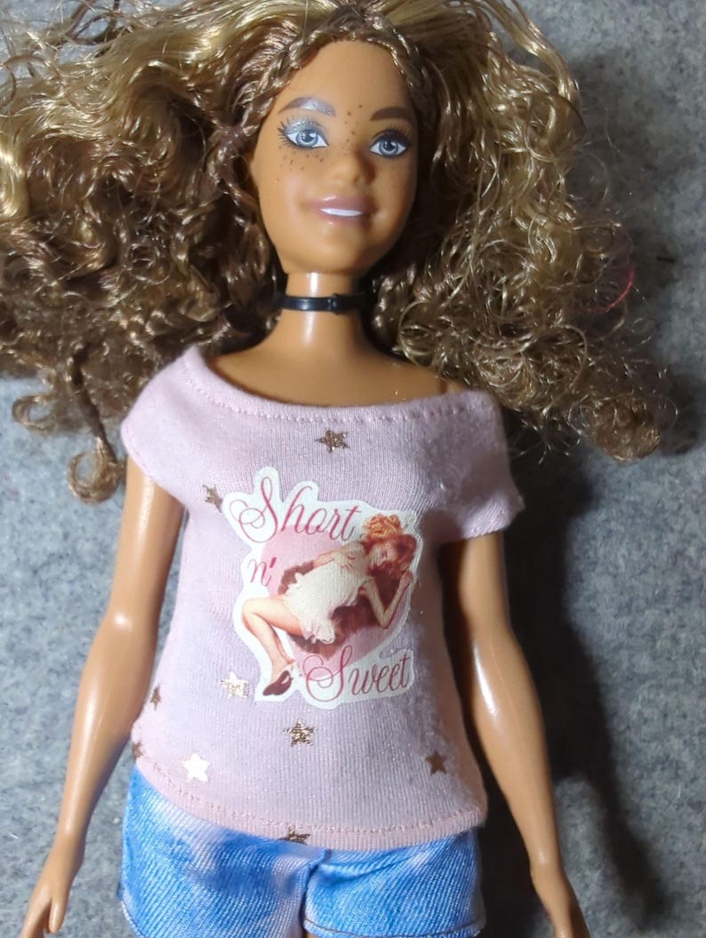 May include: A doll with curly blonde hair, blue eyes, and freckles, wearing a pink off-the-shoulder t-shirt with the words "Short n' Sweet" and a graphic of a woman. The doll is also wearing blue denim shorts and a black choker.