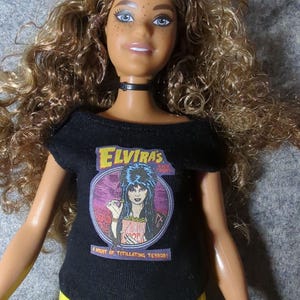 May include: A doll with long, curly brown hair and blue eyes, wearing a black t-shirt with the text "Elvira's" and an image of Elvira, and yellow pants. The shirt also says "A Night of Titillating Terror!" The doll has a black choker.