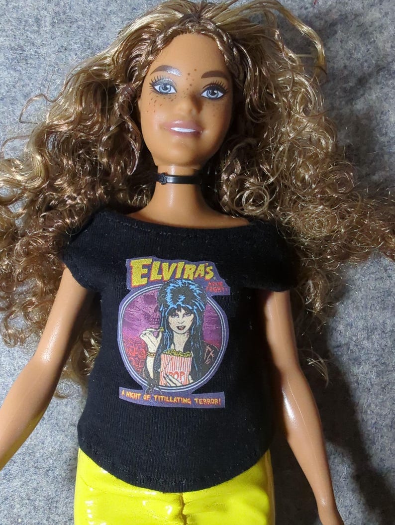 May include: A doll with long, curly brown hair and blue eyes, wearing a black t-shirt with the text "Elvira's" and an image of Elvira, and yellow pants. The shirt also says "A Night of Titillating Terror!" The doll has a black choker.