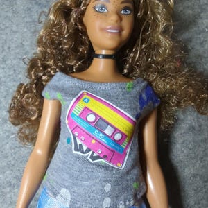 May include: A doll with curly brown hair and blue eyes wearing a gray t-shirt with a cassette tape graphic in pink, yellow, and blue. The doll is also wearing blue denim shorts and a black choker.