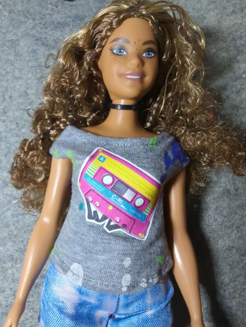 May include: A doll with curly brown hair and blue eyes wearing a gray t-shirt with a cassette tape graphic in pink, yellow, and blue. The doll is also wearing blue denim shorts and a black choker.