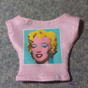 May include: A pink doll-sized t-shirt featuring a pop art image of Marilyn Monroe. The image has a turquoise background with a pink face, yellow hair, and red lips. The shirt is made of a ribbed fabric.