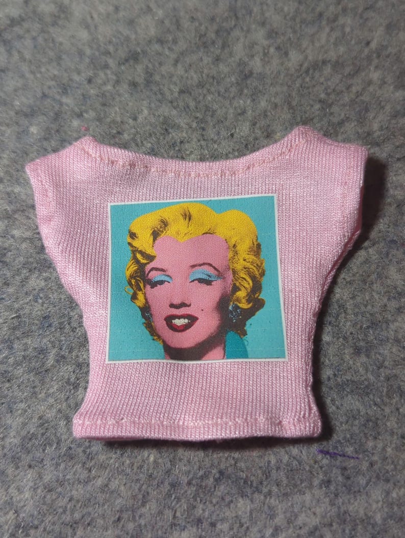 May include: A pink doll-sized t-shirt featuring a pop art image of Marilyn Monroe. The image has a turquoise background with a pink face, yellow hair, and red lips. The shirt is made of a ribbed fabric.