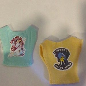 May include: Two doll-sized t-shirts. One is light blue with a unicorn graphic and the word "Majestic". The other is yellow with a graphic of a person under an umbrella and the text "Don't be a Salty Bitch".